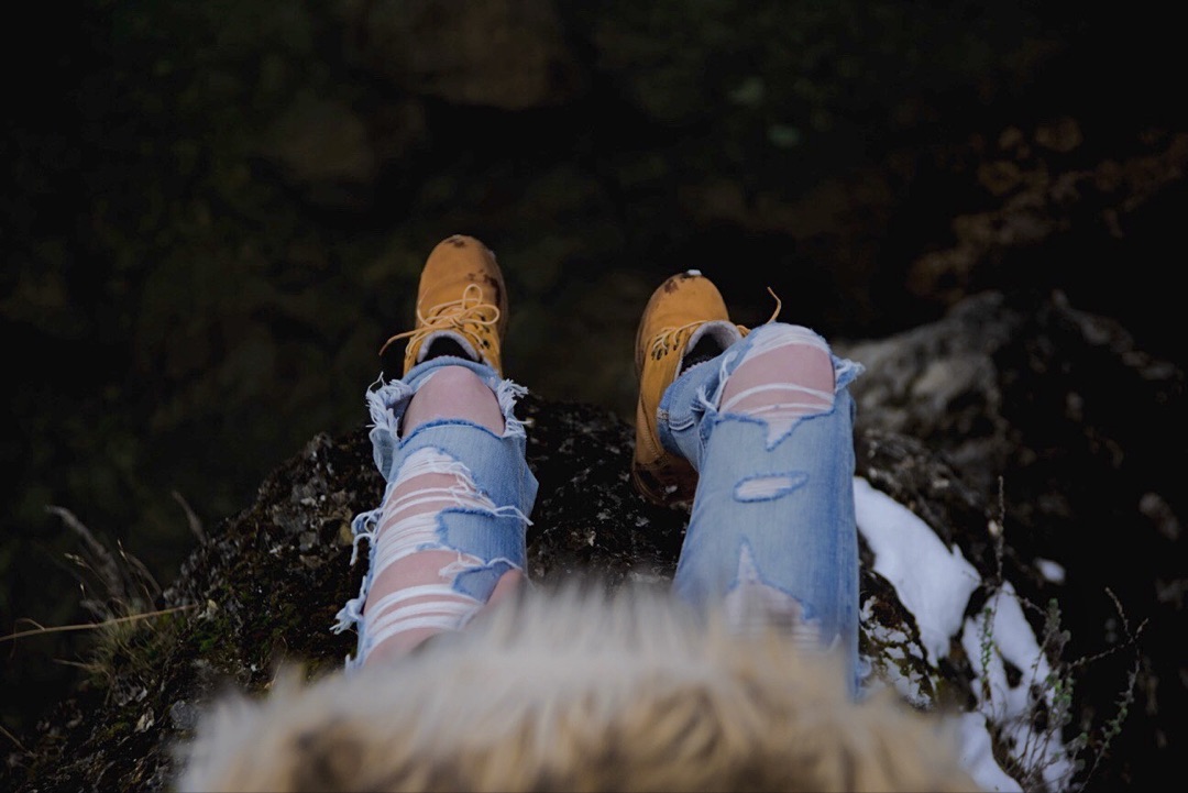Timberland Canada campaign frame — POV looking down over a cliff edge in Kelowna, BC, yellow nubuck boots and ripped jeans on dark moss-covered rock