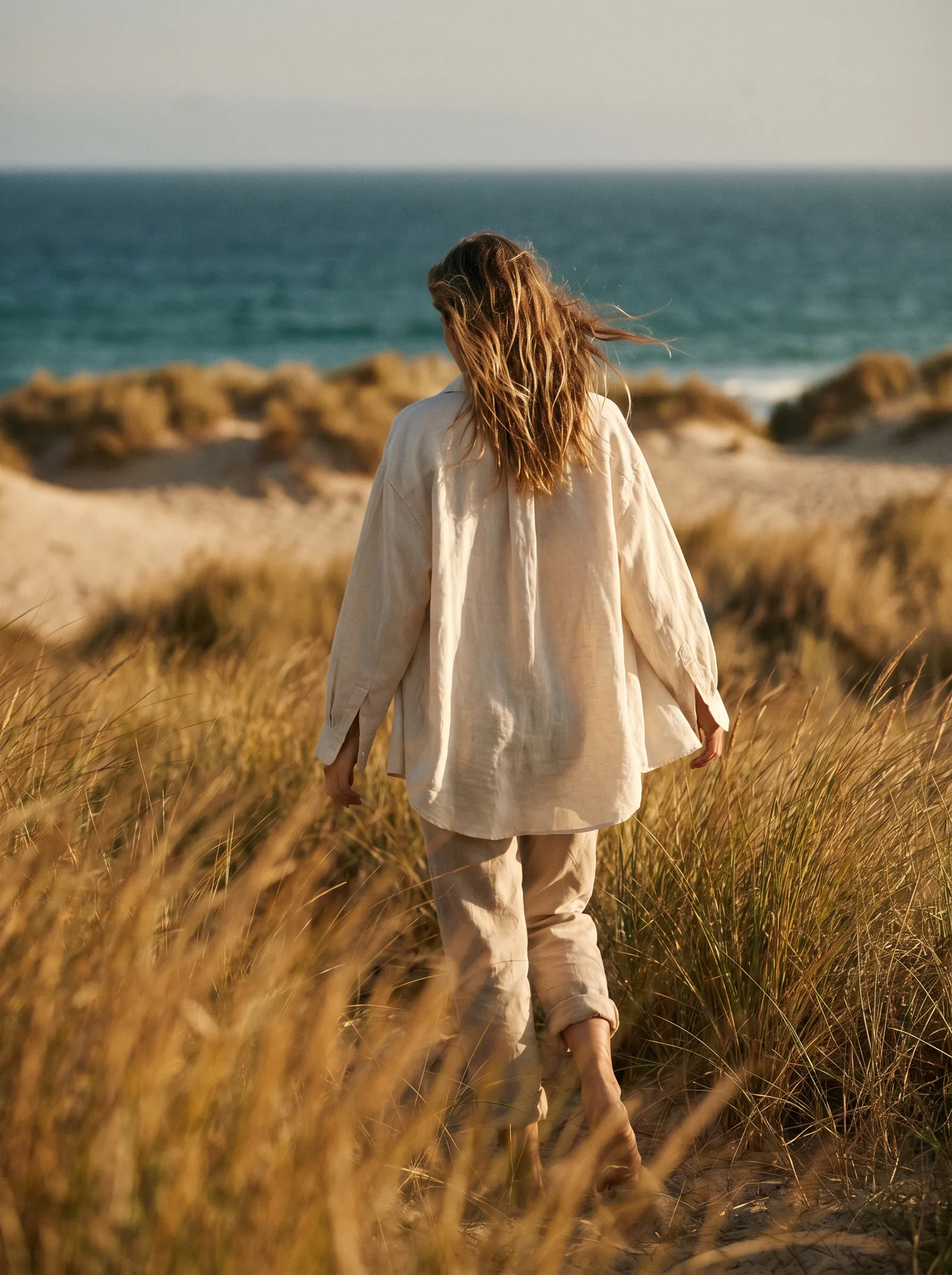 Editorial portrait of a figure walking through coastal grass at golden hour